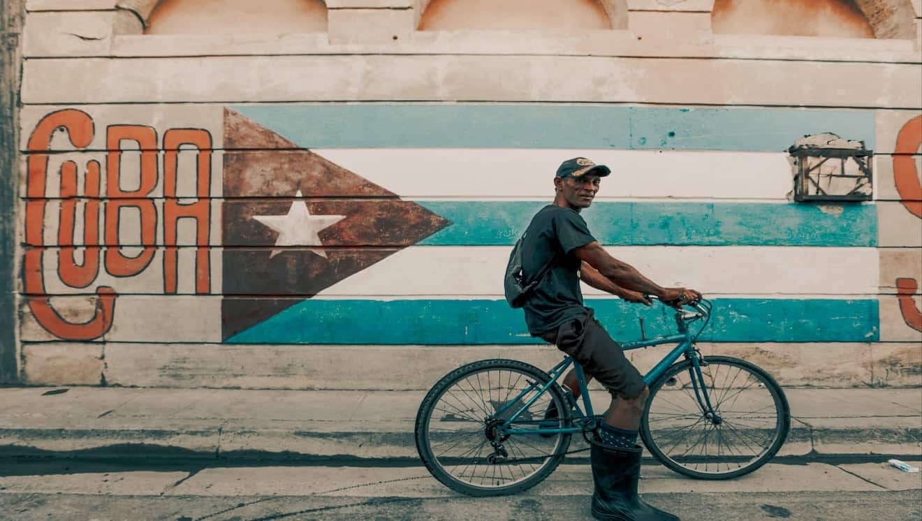 Man cycling through the vibrant streets of Havana, with a Cuban flag mural as backdrop.